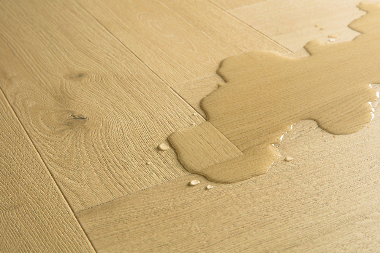 Laminaatti kalanruoto Quick Step Impressive Design Nutmeg Oak, lämmin keskiruskea tammen sävy ja tasainen lajitelma, vesilätäkkö osoittamassa vedenkestävyyttä