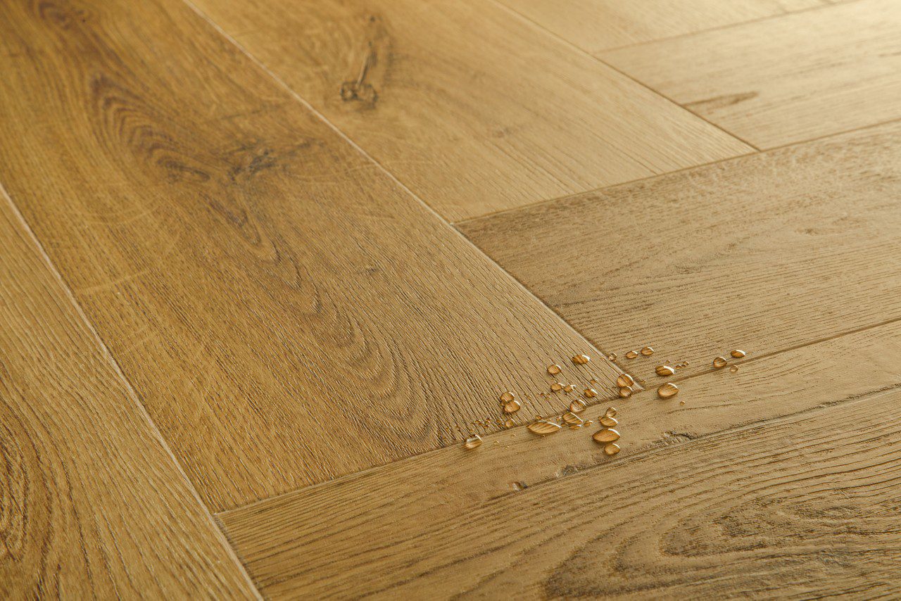 Vinyylilankku kalanruoto Quick Step Ciro Botanic Smoked Oak – tummanruskea, hienovaraisesti harjattu tammen sävy, jossa on lämmin ja luonnollinen ilme. Moderni kalanruotokuvio korostaa puunsyiden kauneutta ja tuo sisustukseen ajattoman tyylin. Lattialla näkyvät vesipisarat korostavat vinyyliparketin täydellistä vedenkestävyyttä.