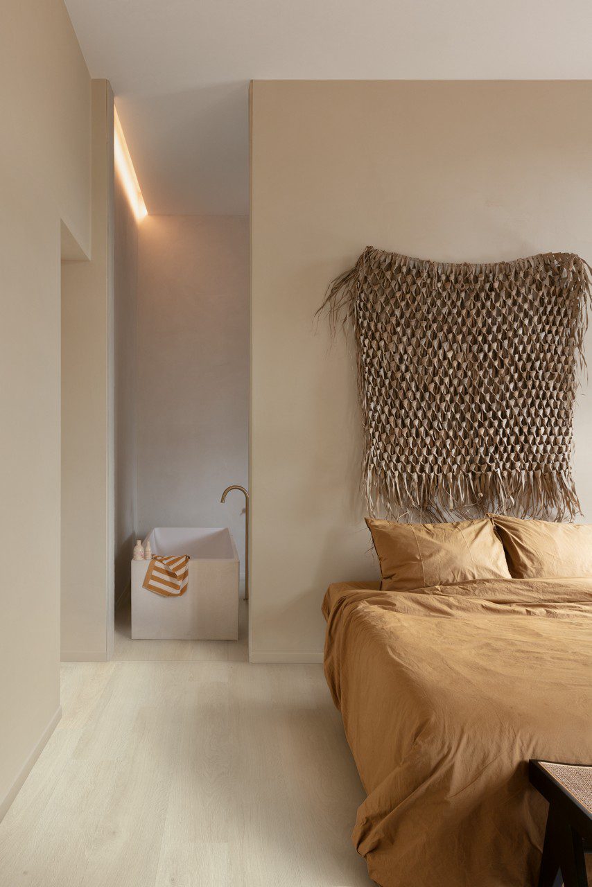 Klickvinyl Quick Step Bloom Sea Breeze Oak Light im Schlafzimmer verlegt, sehr heller Eicheton und Dielendesign, Bett mit schöner hellbrauner Bettwäsche, im Hintergrund ein Badezimmer mit freistehender, eckiger Design-Badewanne.