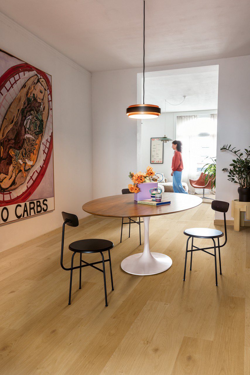 LVT floor Quick-Step Coast Oak Honey installed on ling room floor. Table, stools and a woman walking on the floor.