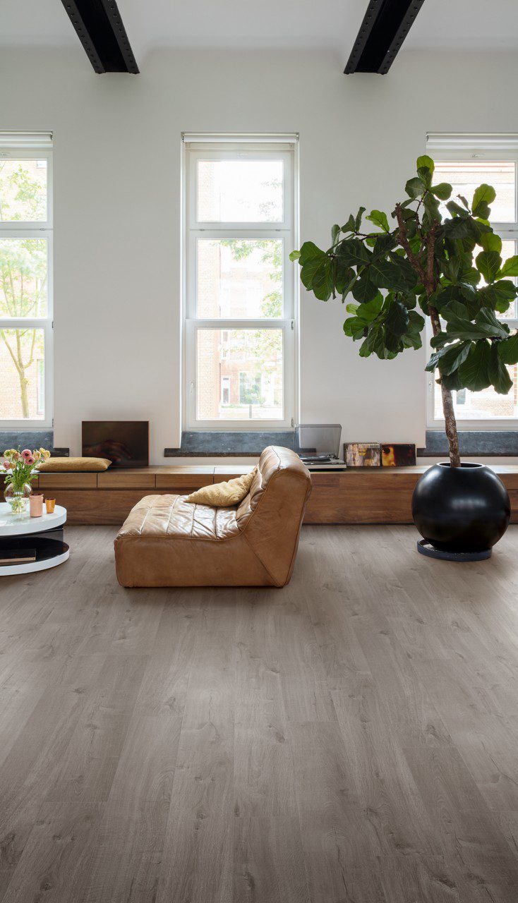 LVT parkett Quick-Step Bloom Cotton Oak Cozy Grey paigaldatud elutoa põrandale. Pruunist nahast dugitool, diivanilaud lilledega, palmipuu, pikk kummut akende all.