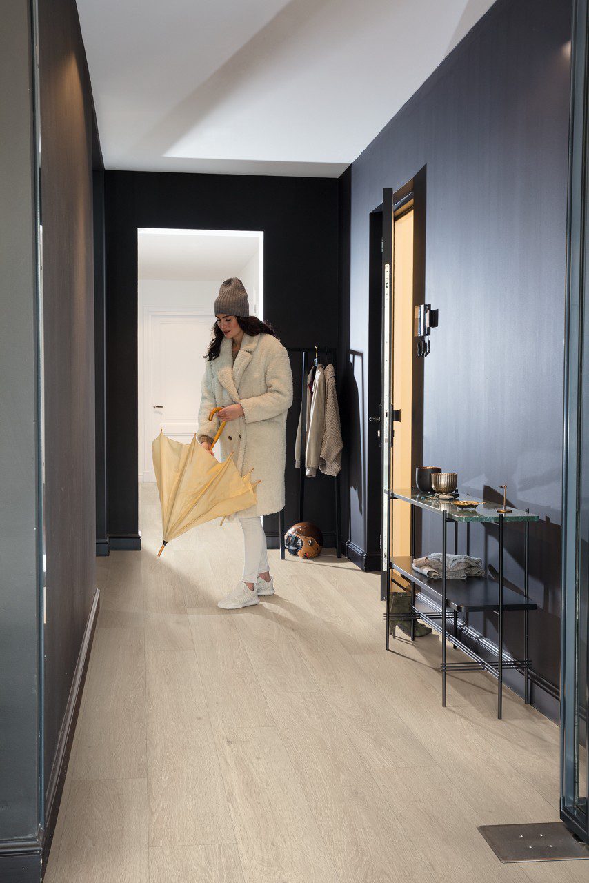 LVT põrand Quick-Step Bloom Sea Breeze Oak Beige paigaldatud koridori põrandale. Naine sisenes tuppa märja vihmavarjuga.