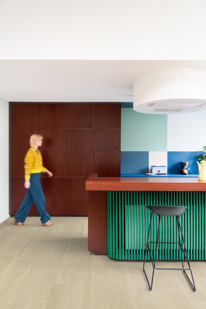 LVT Quick-Step Bloom Sea Breeze Oak Beige paigaldatud köögi põrandale. Naine kõndimas, roheline ja pruun köögimööbel.