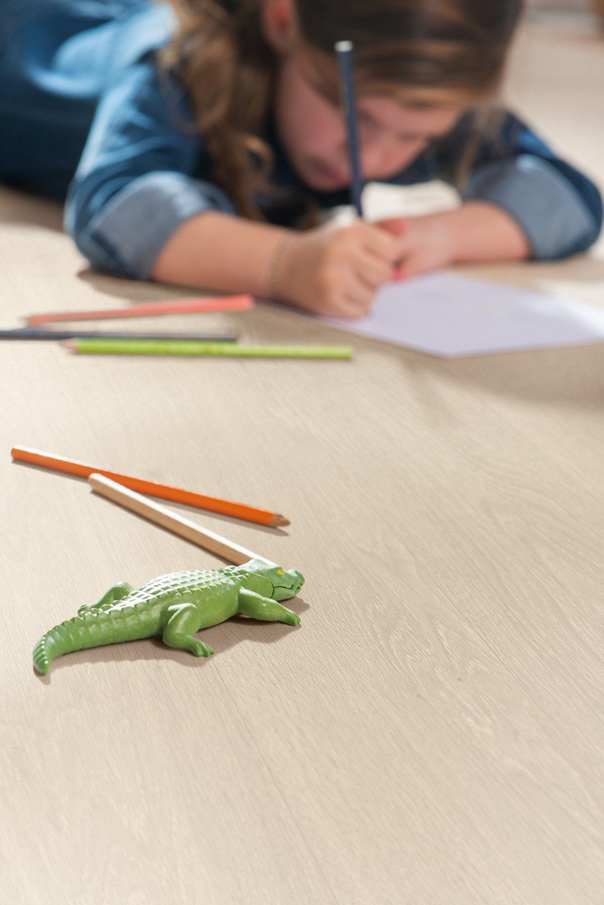 Vinüülparkett Quick-Step Bloom Sea Breeze Oak Beige paigaldatud lastetoa põrandale. Mänguasi krokodill, pliiatsid, väike tüdruk lamab kõhuli põrandal ja joonistab.