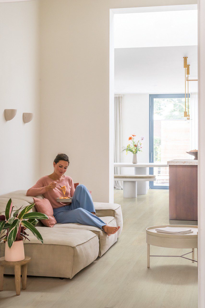 Vinüülparkett Quick-Step Bloom Sea Breeze Tamm Light paigaldatud elutoa põrandale. Naine istub diivanil ja sööb pastat. Potilill, diivanilaud, tagaplaanil köök.