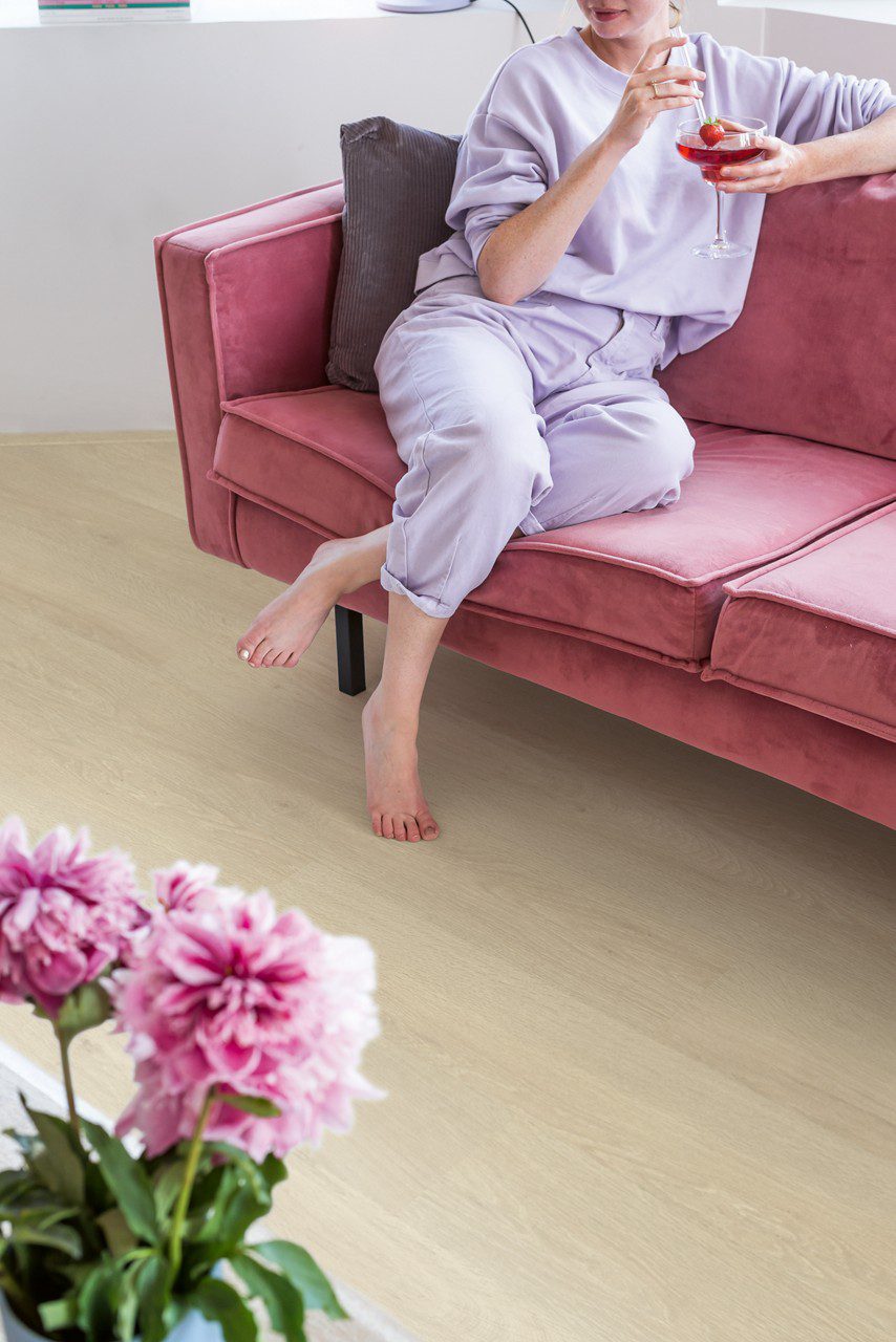 Vinyl flooring Quick-Step Bloom Sea Breeze Oak Beige installed on living room floor. Woman sitting barefoot on a pink sofa and drinking cocktail. Pink flowers.