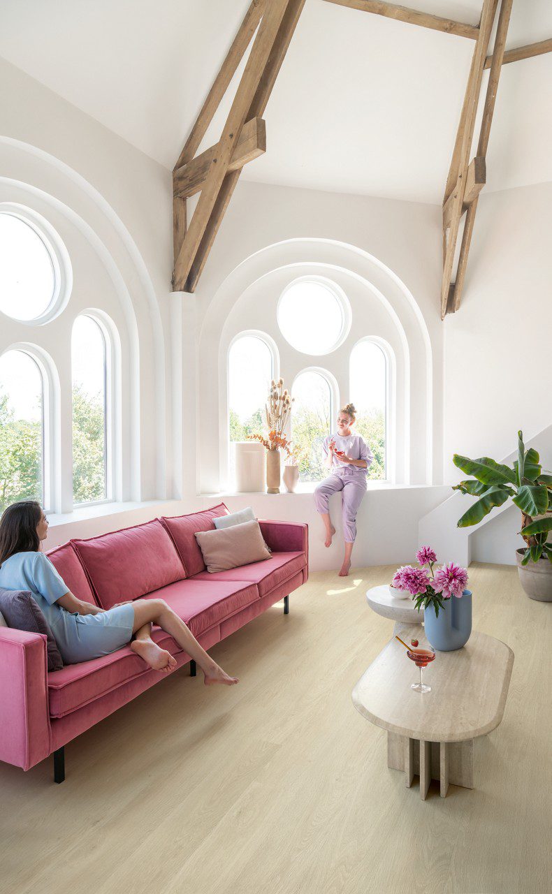 Vinylboden Quick Step Bloom Sea Breeze Eiche Beige im Wohnzimmer verlegt, zwei Frauen unterhalten sich, eine liegt auf einem rosa Sofa, die andere sitzt auf der Fensterbank.