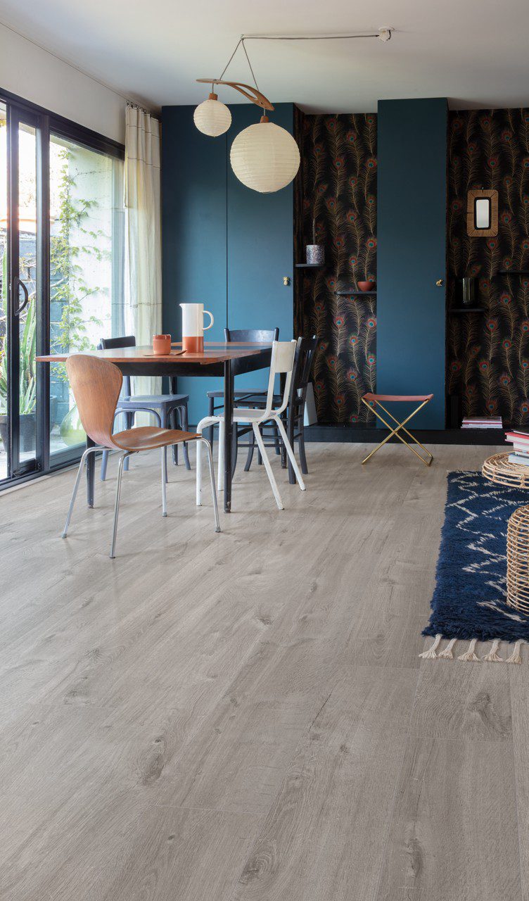 Vinyylilankku Quick Step Bloom Cotton Oak Cozy Grey asennettuna ruokailutilan lattialle, pehmeä lämminharmaa tammen sävy ja luonnollinen lajittelu, lankkudesign, siniset seinäkaapit, ruokapöytä ja eri väriset tuolit.