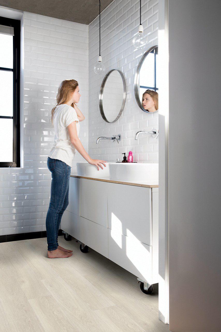 Vinyylilankku Quick Step Bloom Sea Breeze Tammi Light asennettuna kylpyhuoneen lattialle, nainen laittaa hiuksiaan peilin edessä, seinillä retrohenkiset laatat.