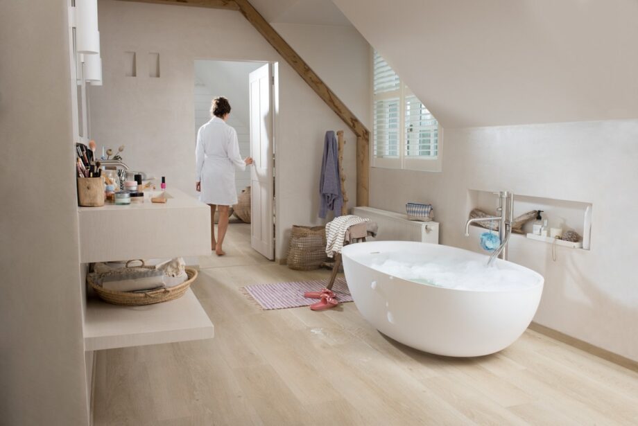 vinyylilankku-vaalea-quick-step-bloom-sea-breeze-oak-beige Vaalea vinyylilankku Quick Step Bloom Sea Breeze Oak Beige asennettuna kylpyhuoneen lattialle, erillinen valkoinen kylpyamme ja beige kylpyhuonekalusteet, nainen kävelee märin jaloin kylpytakissa lattialla.