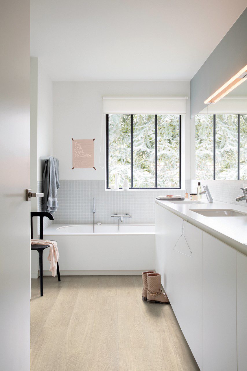 Vinyylilattia Quick Step Bloom Sea Breeze Tammi Beige asennettuna kylpyhuoneen lattialle, valkoinen kylpyamme ja valkoiset kylpyhuonekalusteet.