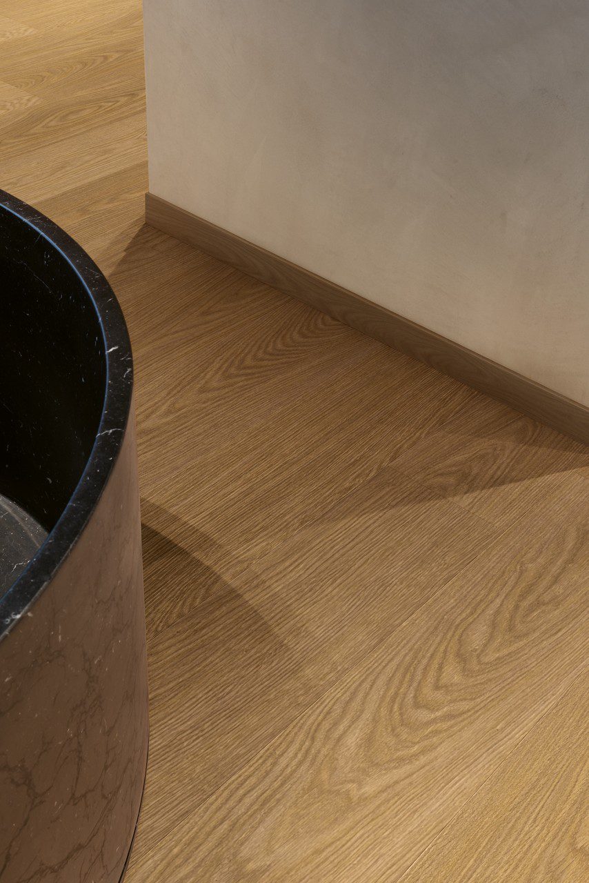 Vinyylilattia Quick Step Blos Cocoa Tammi asennettuna kylpyhuoneen lattialle, tumma kaakaonruskea tammen sävy ja lankkulattia tammi, samansävyiset jalkalistat ja musta luonnonkivinen kylpyamme.