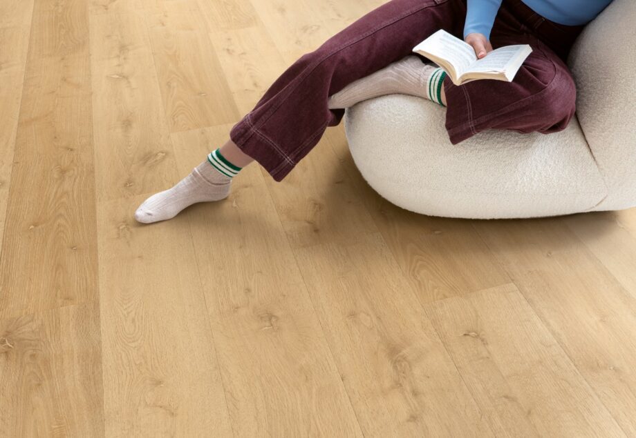 Виниловый ламинат Quick Step Bloom Elegant Oak Natural уложен на полу в гостиной, женщина сидит в удобном кресле и читает книгу.