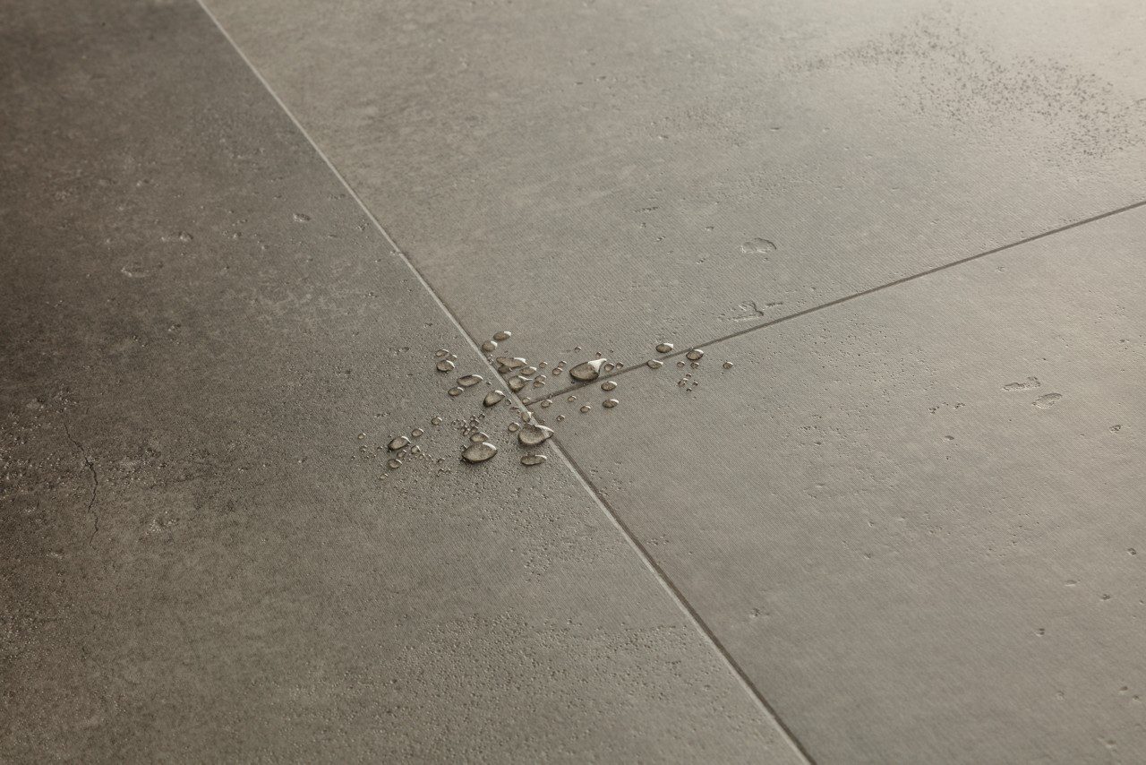 LVT parkett Quick-Step Illume Cloudy concrete tumehalli tooni ja betooni disainiga. Põrandal on veepiisad viitamaks antud LVT parketi veekindlusele.