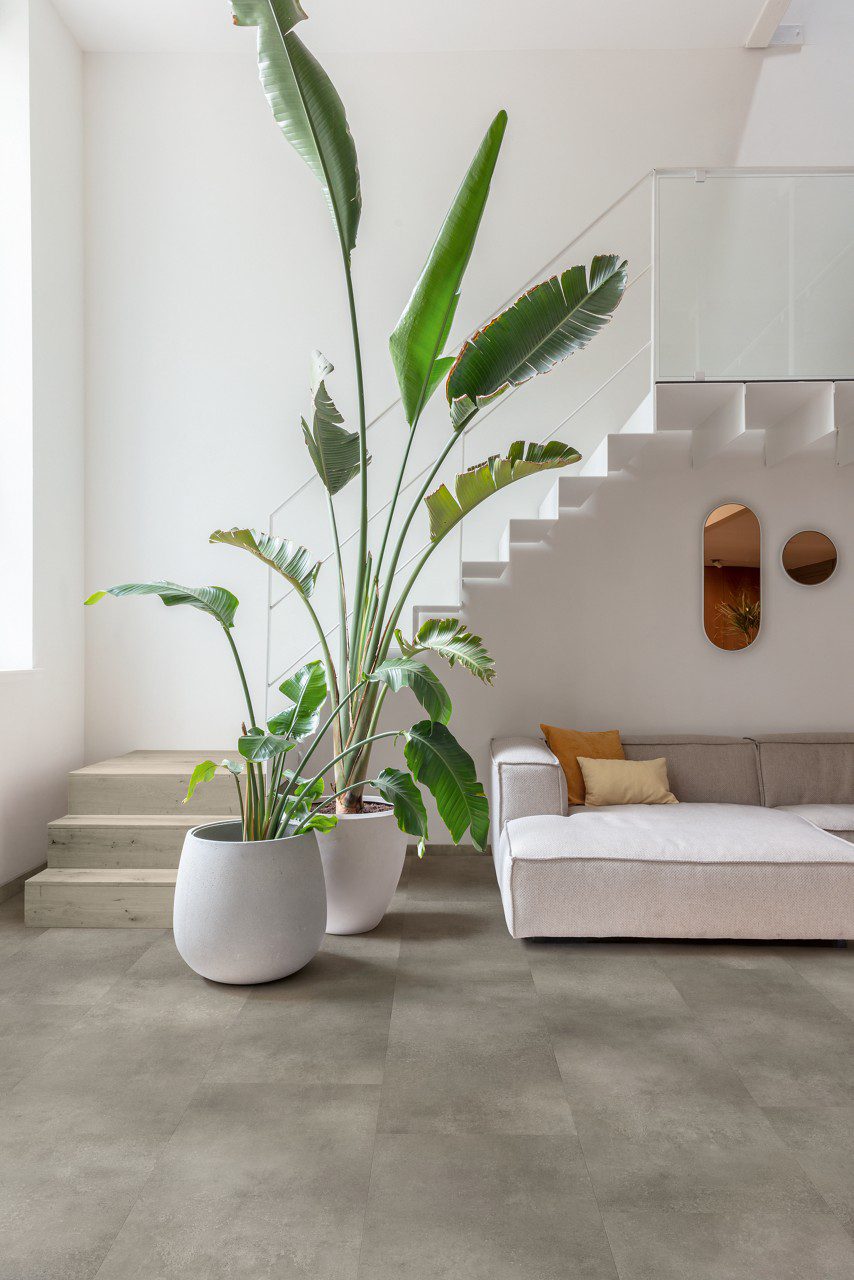 Vinyl flooring tiles Quick.Step Illume Cloudy concrete installed on a living room floor. Same color and design flooring skirtings. Sofa, green plants and stairs.