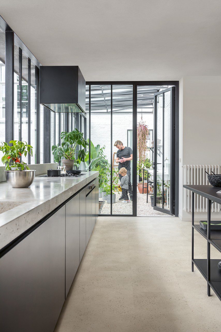 Vinyl tiles Quick-Step Illume Oyster concete installed on a kitchen floor. Kitchen furniture, opened door to summer garden and father and son are watering plants there.