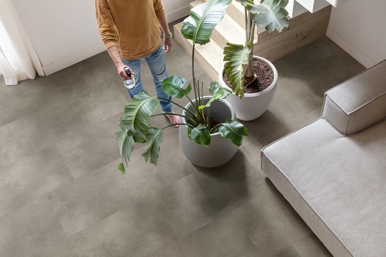 Vinylboden in Betonoptik Quick-Step Illume Cloudy Concrete, verlegt auf dem Wohnzimmerboden – Sofa, Zimmerpflanzen im Topf und eine Treppe; ein Mann besprüht die Pflanzen mit Wasser und steht barfuß auf dem Boden.