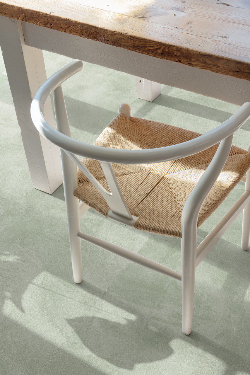 Vinylboden in Betonoptik Quick-Step Illume Soft Sage, verlegt auf dem Küchenboden – rustikaler Esstisch und Stuhl in Nahaufnahme; heller, sanfter Salbeigrün-Ton mit dezentem, steinartigem Design.
