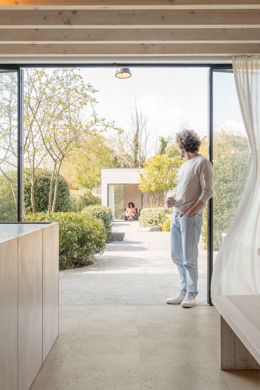 Vinyylilattia Quick-Step Illume Oyster Concrete asennettuna keittiön lattialle – lattiasta kattoon ulottuvat liukuovet ovat auki, mies katsoo kahvikuppi kädessä kesäiseen puutarhaan; taustalla näkyy avoimella liukuovella varustettu piharakennus, jossa nainen istuu ja katsoo takaisin. Vaalea, lämmin greige/oyster-sävy ja hillitty betonidesign.