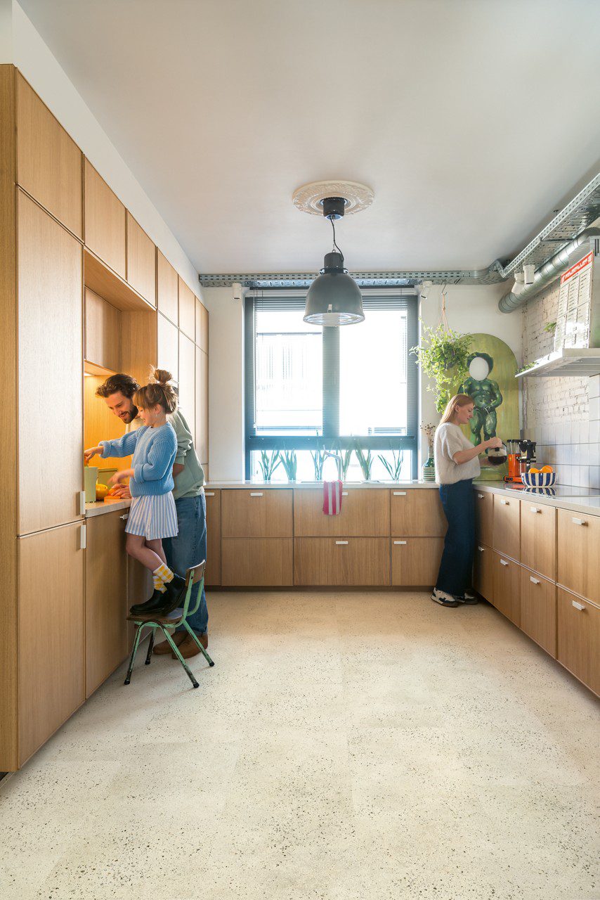 Vinyylilattia Quick-Step Illume Pebble Concrete asennettuna keittiön lattialle – nainen kaataa kahvia, ja isä sekä tytär puristavat yhdessä appelsiinimehua; vaalea greige/betonisävy ja hillitty betonikividesign.