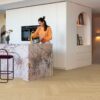 Fischgrat Vinylboden Quick Step Ciro Pure Oak Blush, verlegt auf dem Küchenboden, mit weißen Küchenmöbeln, einer Kücheninsel aus rosafarbenem Marmor mit einer goldenen Armatur, aus der eine Frau Wasser in einen verchromten Kochtopf laufen lässt.