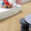 Herringbone vinyl floors Quick Step Ciro Pure Oak Blush, installed on the living room floor, a woman sitting on a white sofa browsing a magazine, a table, and a pink pedestal.