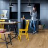 LVT parkett Quick-Step Bloom Cotton Oak Natural paigaldatud köögi põrandale. Musta fassaadiga köögimööbel, söögilaud kollaste ja mustade toolidega. Naine sööb võileiba.
