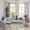 lvt-parkett-quick-step-bloom-cotton-oak-white-blush LVT parkett Quick-Step Bloom Cotton Oak White Blush paigaldatud elutoa põrandale. Ema sulgeb puidust aknakatteid ja lapsed vaatavad seda valgelt nahast diivanilt.