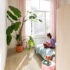 Quick Step Bloom Brushed Oak Beige vinyl floor installed in a bedroom, with a palm plant, pink curtains, and a woman reading a book on the bed.
