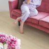 vinyl-flooring-quick-step-bloom-sea-breeze-oak-beige Vinyl flooring Quick-Step Bloom Sea Breeze Oak Beige installed on living room floor. Woman sitting barefoot on a pink sofa and drinking cocktail. Pink flowers.