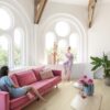 vinylböden-quick-step-bloom-sea-breeze-eiche-beige Vinylboden Quick Step Bloom Sea Breeze Eiche Beige im Wohnzimmer verlegt, zwei Frauen unterhalten sich, eine liegt auf einem rosa Sofa, die andere sitzt auf der Fensterbank.