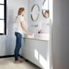 vinyylilankku-quick-step-bloom-sea-breeze-tammi-light Vinyylilankku Quick Step Bloom Sea Breeze Tammi Light asennettuna kylpyhuoneen lattialle, nainen laittaa hiuksiaan peilin edessä, seinillä retrohenkiset laatat.