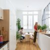 vinyylilankku-quick-step-bloom-urban-black-butt Vinyylilankku Quick Step Bloom Urban Blackbutt asennettuna keittiön lattialle, nainen kastelee keittiötasolla olevaa ruukkukasvia, mies istuu ruokapöydän ääressä ja katsoo puhelinta.