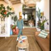 Vinyylilankku Quick Step Bloom Wild Spotted Gum asennettuna olohuoneen lattialle ja matalalle välitasanteen portaalle, nainen kävelee portaita ylös ja koira odottaa ylhäällä.