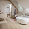 vinyylilankku-vaalea-quick-step-bloom-sea-breeze-oak-beige Vaalea vinyylilankku Quick Step Bloom Sea Breeze Oak Beige asennettuna kylpyhuoneen lattialle, erillinen valkoinen kylpyamme ja beige kylpyhuonekalusteet, nainen kävelee märin jaloin kylpytakissa lattialla.