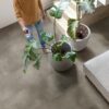 vinylboden-beton-quick-step-illume-cloudy-concrete Vinylboden in Betonoptik Quick-Step Illume Cloudy Concrete, verlegt auf dem Wohnzimmerboden – Sofa, Zimmerpflanzen im Topf und eine Treppe; ein Mann besprüht die Pflanzen mit Wasser und steht barfuß auf dem Boden.
