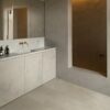 vinylboden-beton-quick-step-illume-sandstone-concrete Vinylboden in Betonoptik Quick-Step Illume Sandstone Concrete, verlegt auf dem Badezimmerboden – graues Badmöbel mit grauer Waschtischplatte und Aufsatzbecken, drei Spiegelschränke an der Wand sowie eine goldene Wandarmatur; warmer Greige-/Sandton mit dezentem Betondesign.
