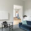 vinylboden-beton-quick-step-illume-soft-sky Vinylboden in Betonoptik Quick-Step Illume Soft Sky, verlegt auf dem Wohnzimmerboden – schwarzer Ledersessel und blaues Sofa; im Hintergrund sind in der Küche ein stehender Vater und ein kleiner Sohn auf einem Dreirad zu sehen.