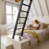 кварц-винил-quick-step-illume-sandstone-concrete Кварц-винил Quick-Step Illume Sandstone Concrete уложен на полу мансардной спальни — женщина лежит в постели и читает книгу; тёплый песочно-бежевый (greige) оттенок и спокойный бетонный дизайн.